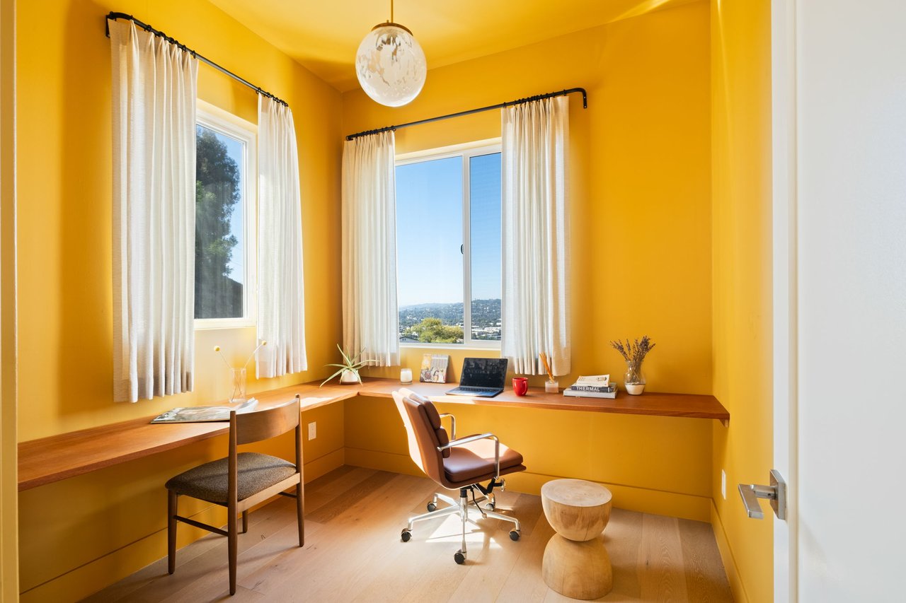 Golden-yellow home office with a wraparound wooden desk, two chairs, and curtained windows.