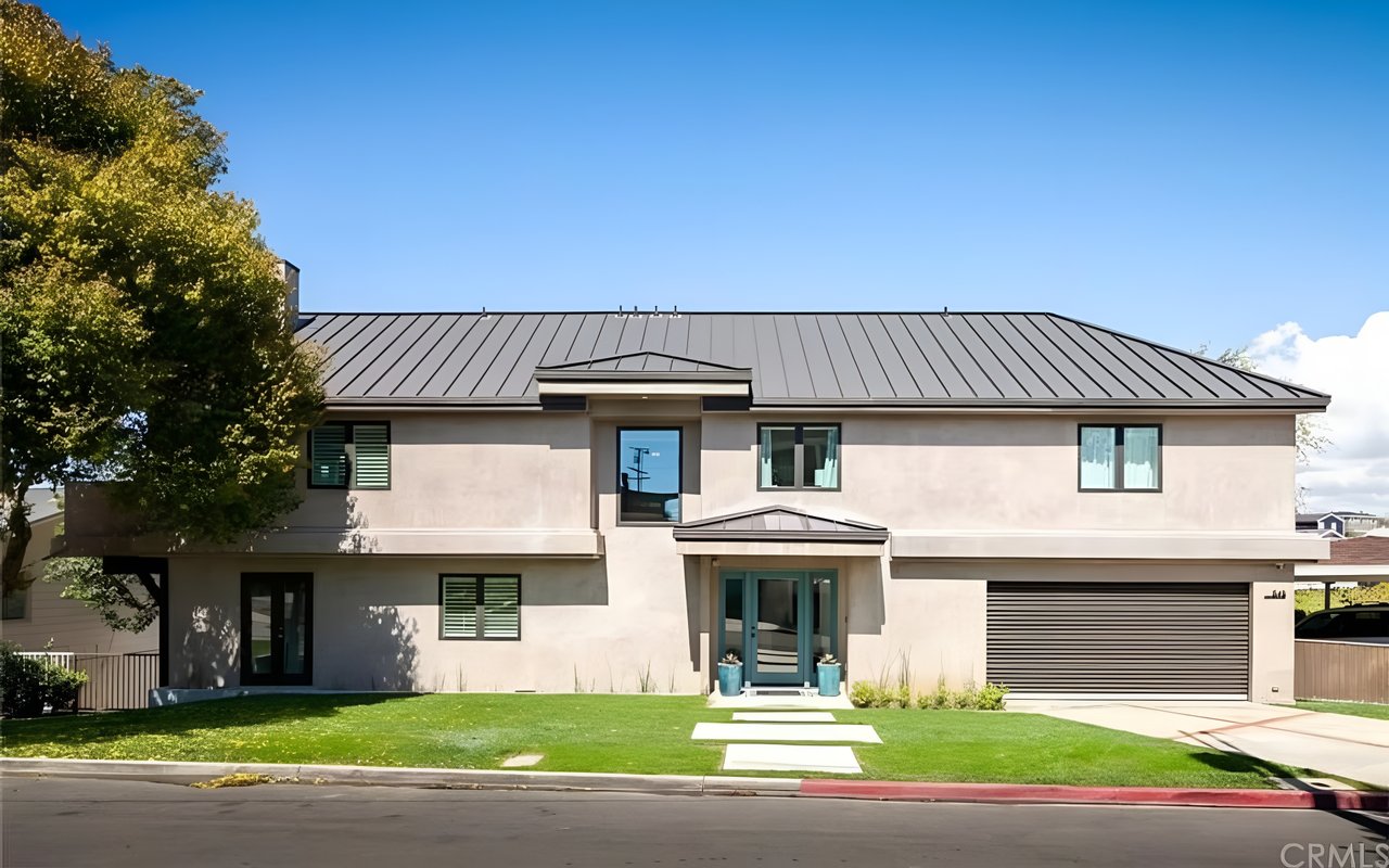 A contemporary two-story beige house with a dark gray metal roof, a modern garage door, and a green lawn under a clear blue sky.