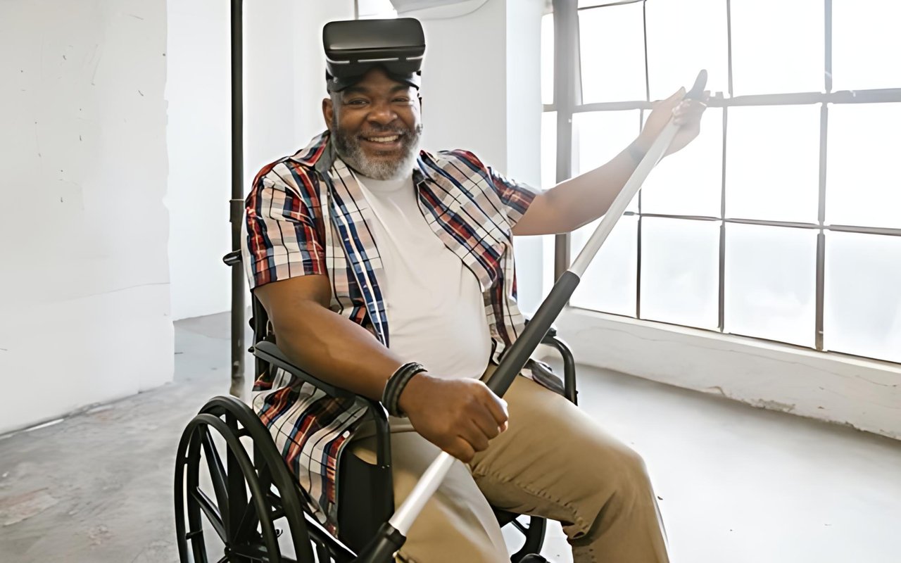 A smiling man seated in a wheelchair holds a long white pole, with a headset resting on his head.