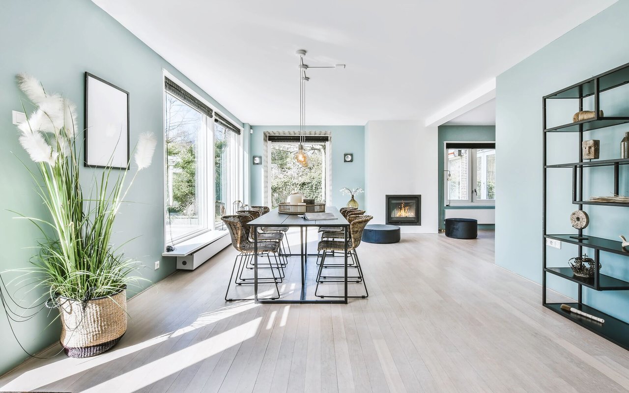 Open-plan dining area with woven chairs, large windows, pale blue walls, and a fireplace.