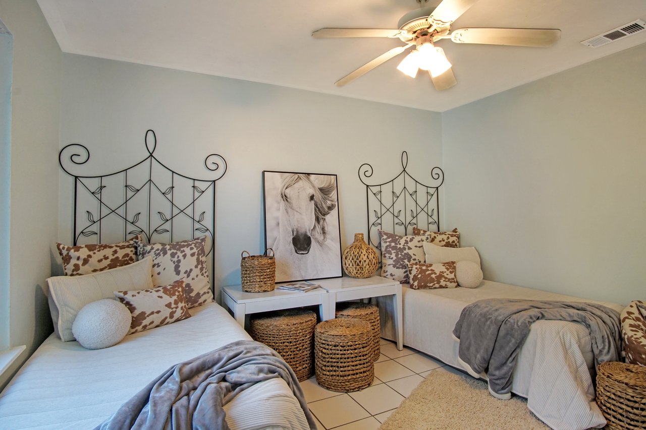 A cozy room with two twin beds featuring ornate metal headboards. Neutral-toned pillows and blankets, wicker baskets, and a horse portrait add warmth.