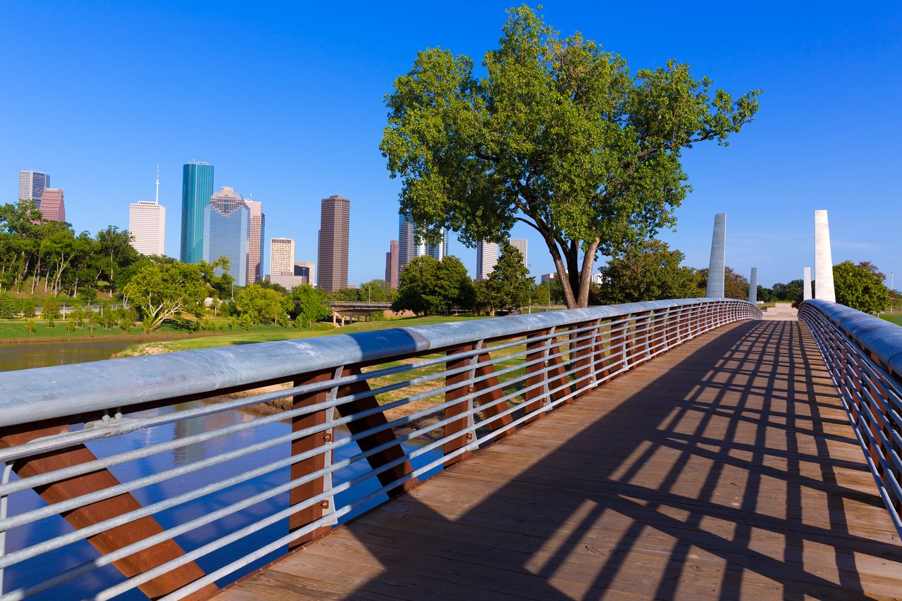 Pedestrian bridge overlooking Houston’s downtown skyline and a tree-lined bayou.