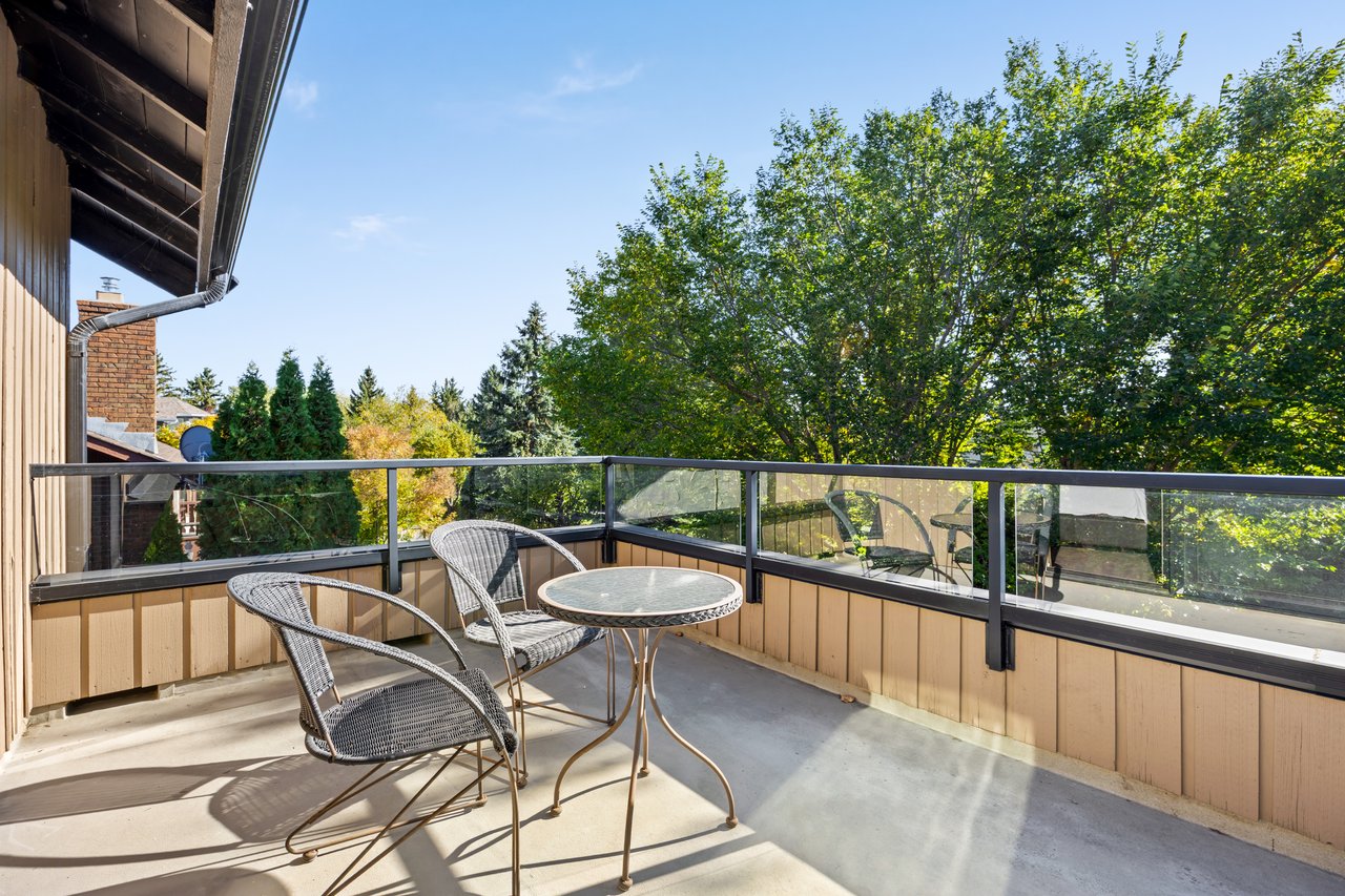 Furnished balcony with glass railings, wicker chairs, and leafy trees beyond.
