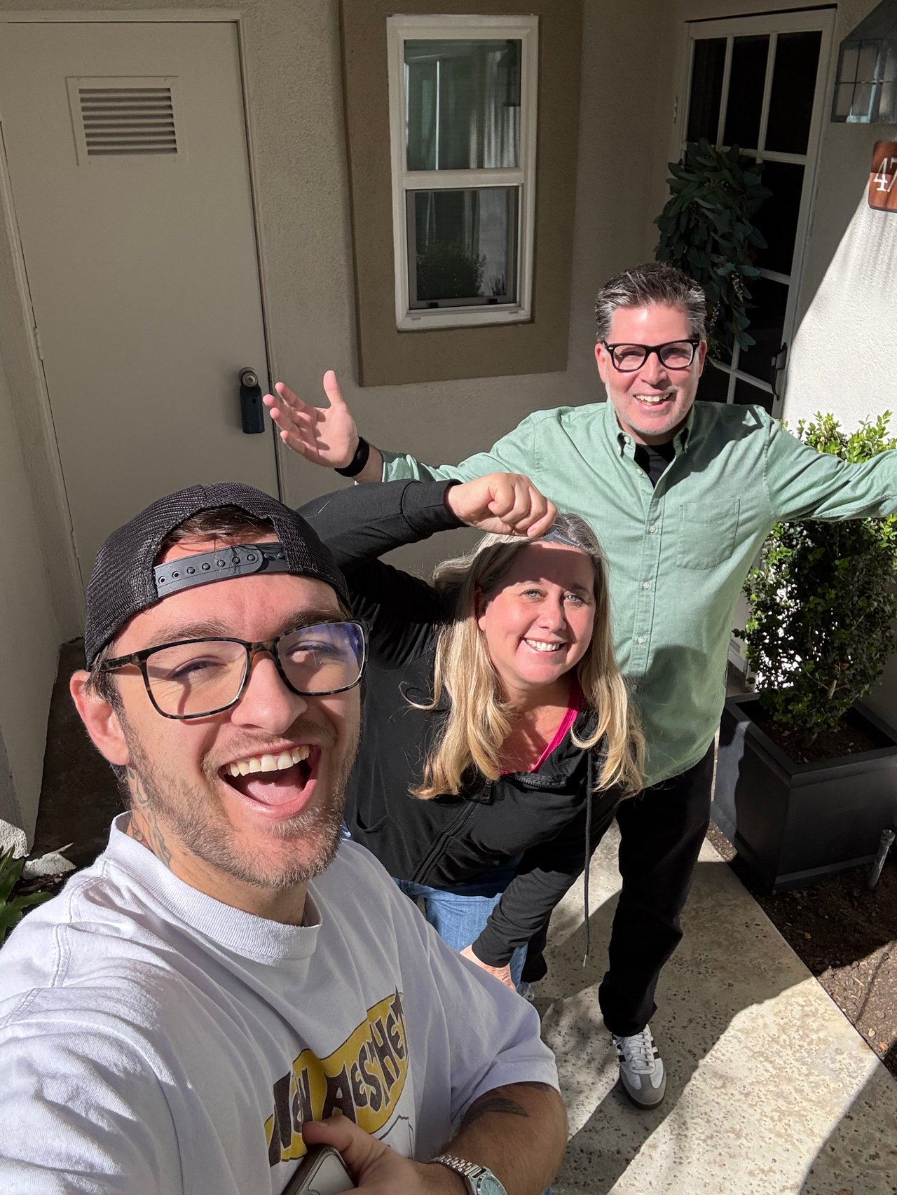 Three smiling people pose for a selfie outside a home.