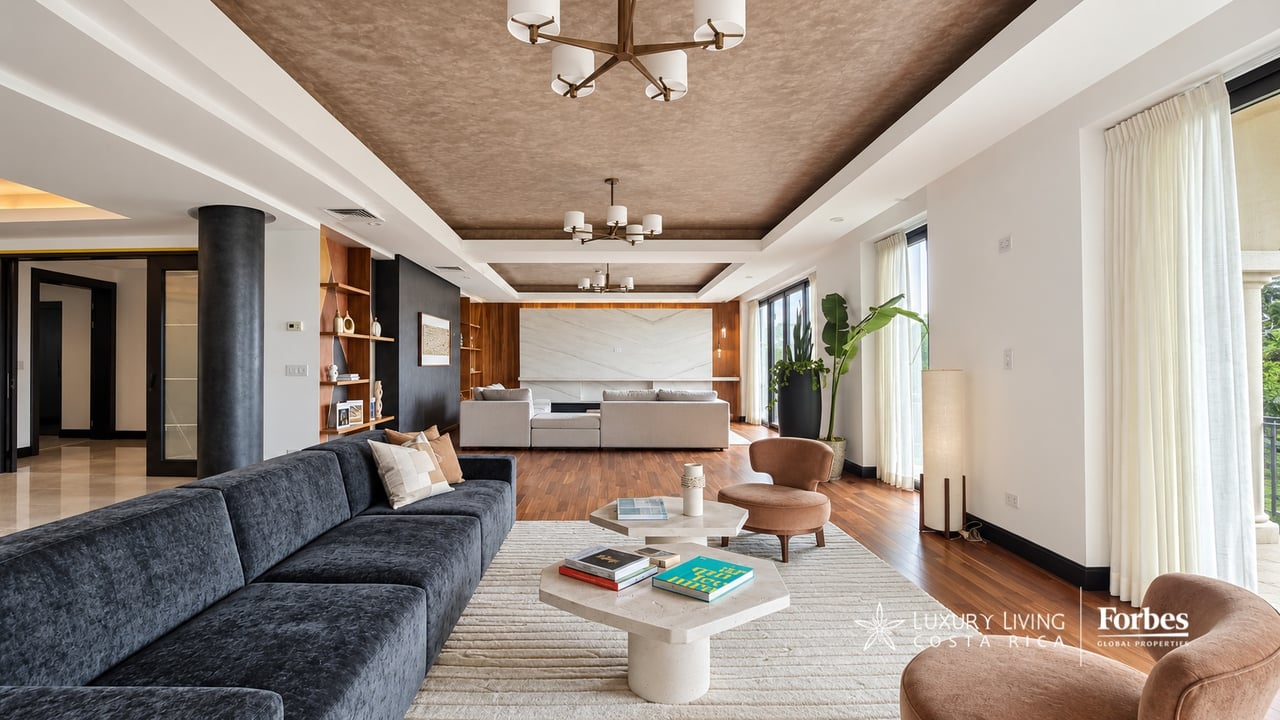 Penthouse living room with charcoal sectional, warm wood floors, sculptural ceiling, and tall curtained windows.