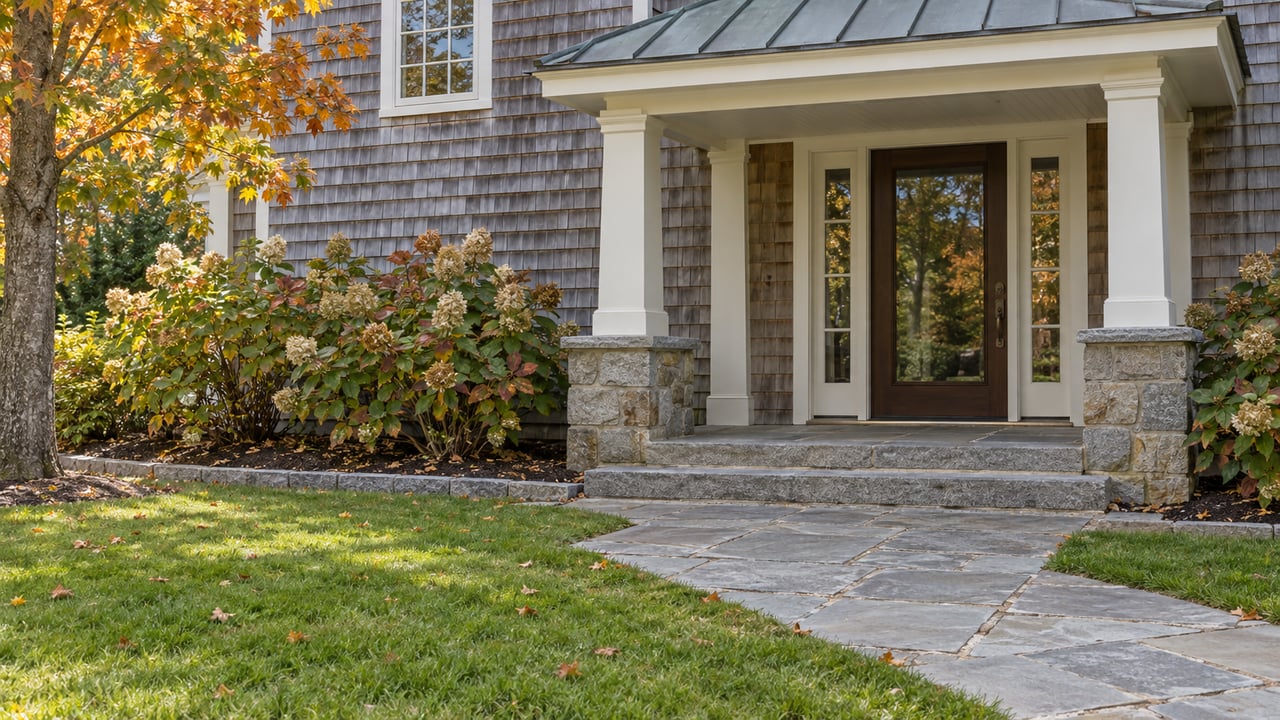 Weathered cedar-shingle home with a covered ivory-trimmed entrance, bluestone approach and early-autumn planting.