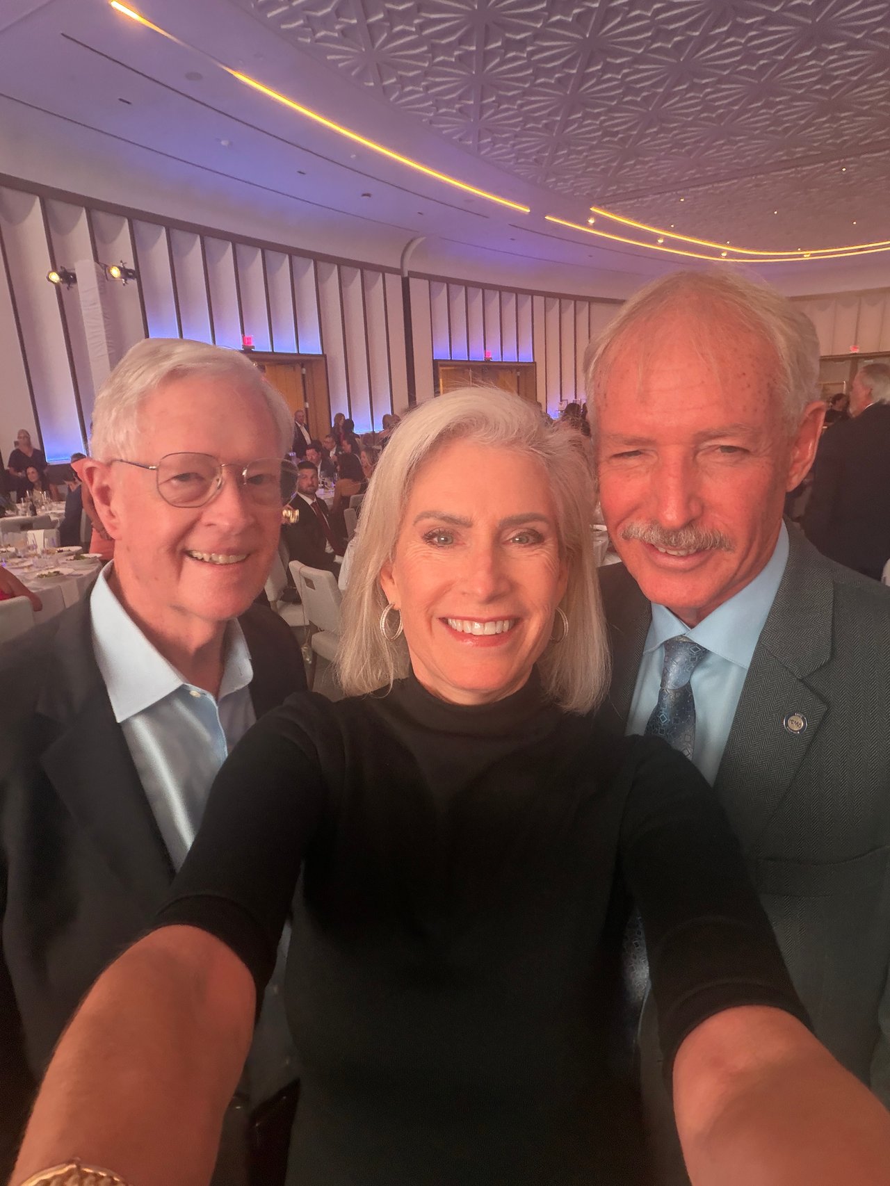 Three adults pose for a selfie at a banquet; the woman wears black and stands between two men in suits.