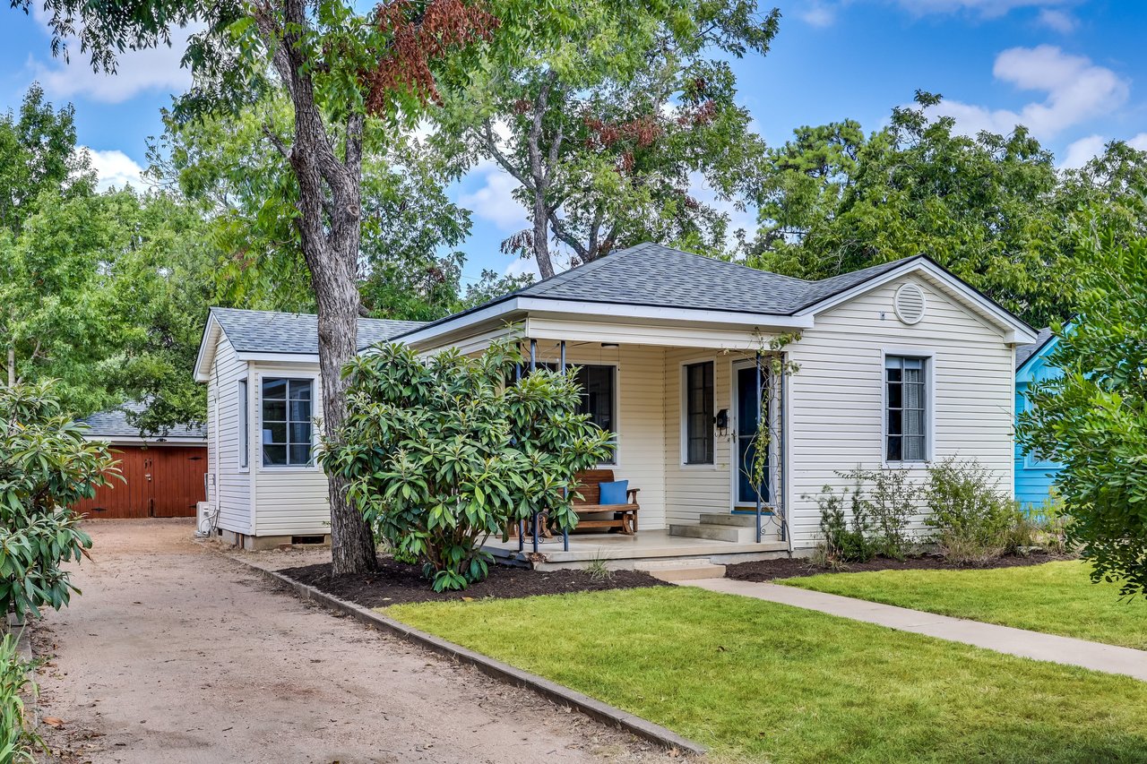 White single-story house with a covered porch, mature trees, green lawn, and gravel driveway.
