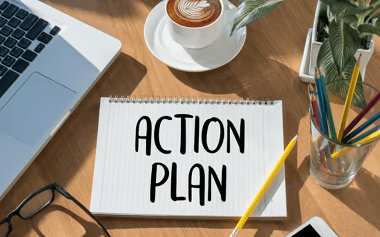 Notebook labeled “ACTION PLAN” on a desk beside a laptop, coffee, pencils, and glasses.