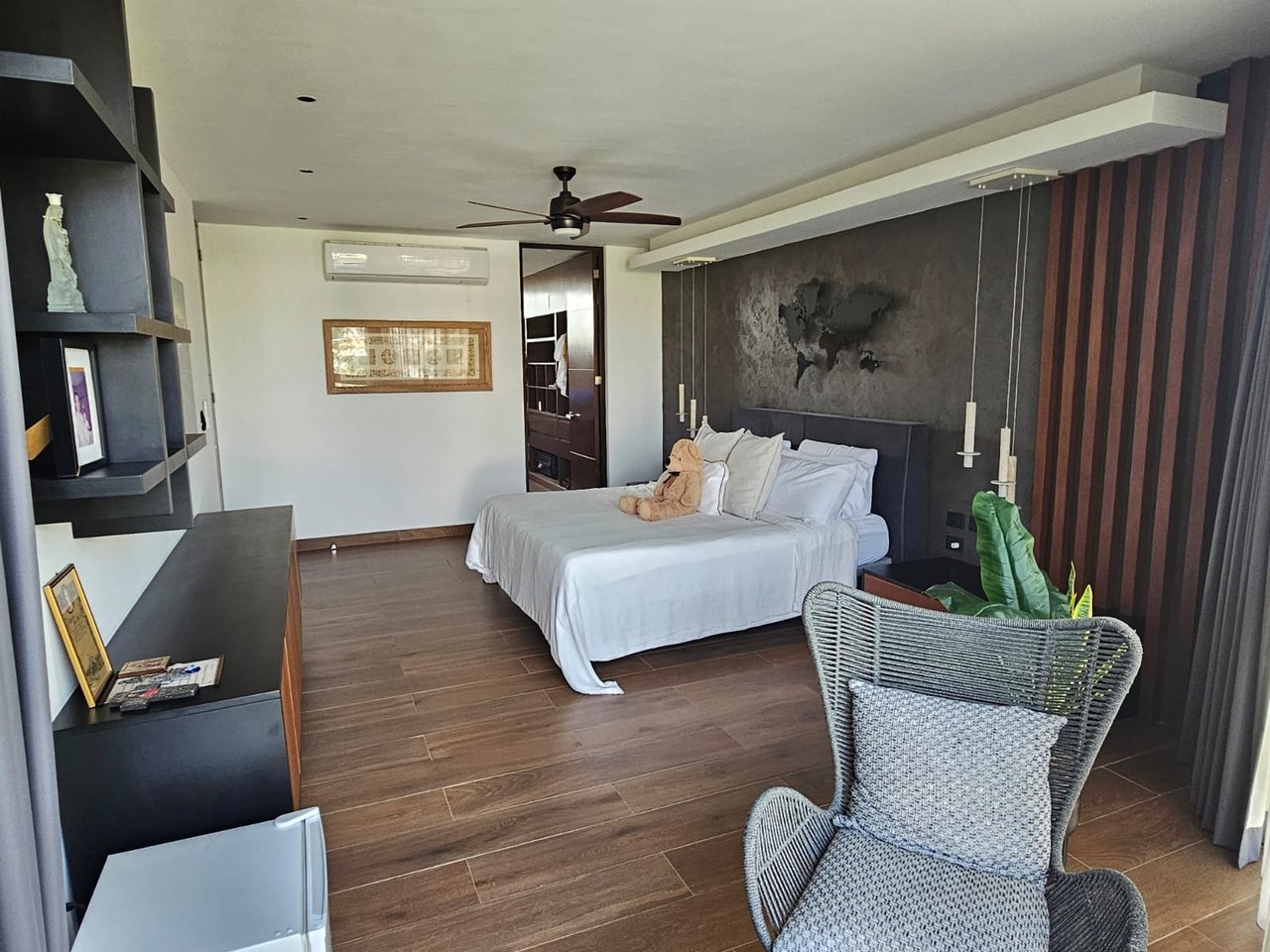 Bedroom with a white bed, dark accent wall, built-in shelving, and woven lounge chair.