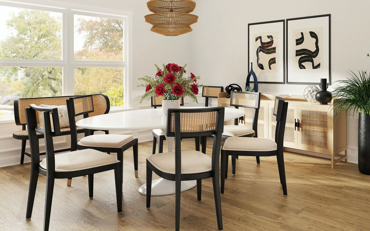 Bright dining room with a white oval table, cane-back chairs, and large windows overlooking trees.