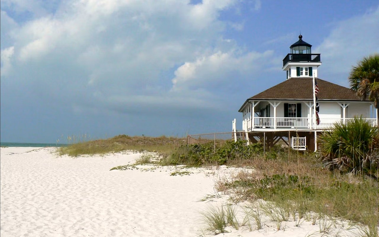 White lighthouse building on stilts beside a sandy beach and coastal dunes.