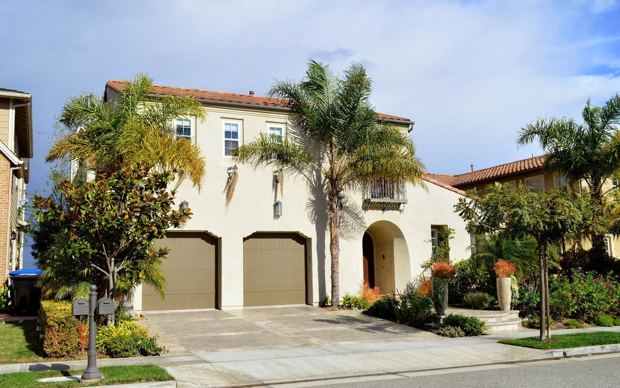 Two-story stucco house with a tiled roof, arched entry, palm trees, and a wide driveway.
