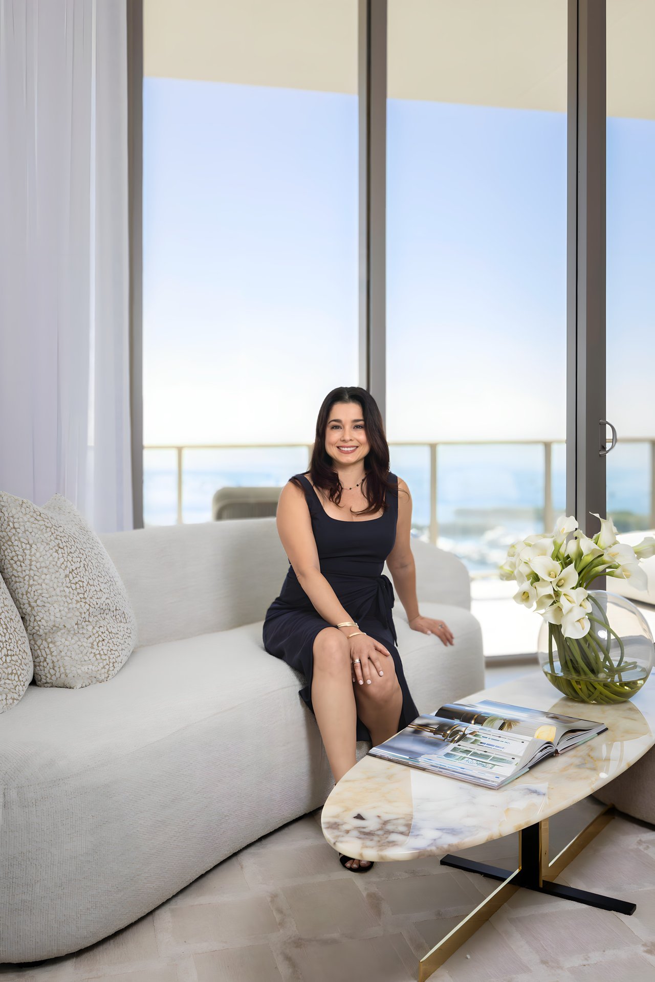 Jenilyn Martinez seated on a white sofa beside large windows and a vase of white flowers.