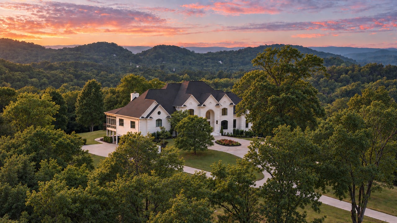 Large white home with a dark roof amid wooded hills at sunset.