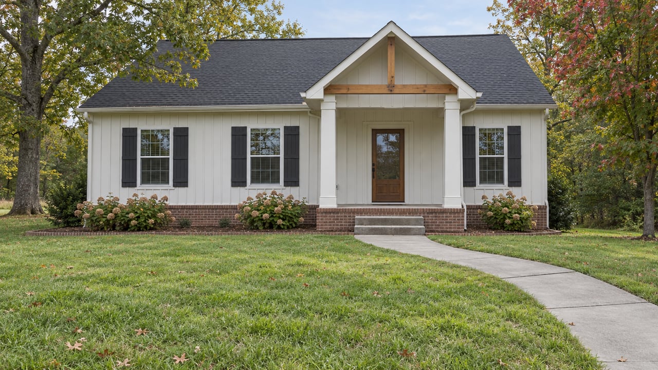 A modest off-white gabled home with a brick foundation and covered porch beyond a deep lawn, framed by mature trees in early autumn.