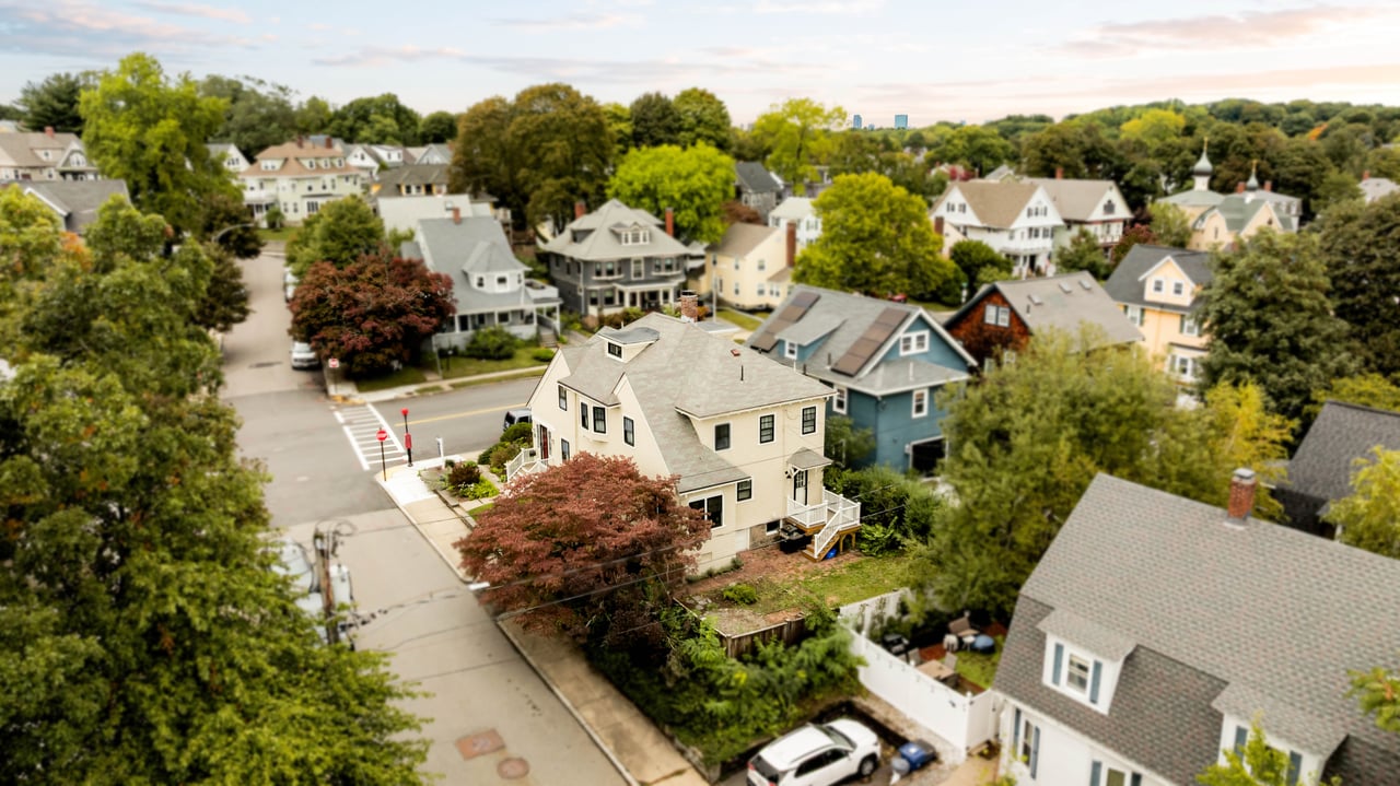 Aerial view of a cream Craftsman home at a leafy residential intersection, beside a blue house.