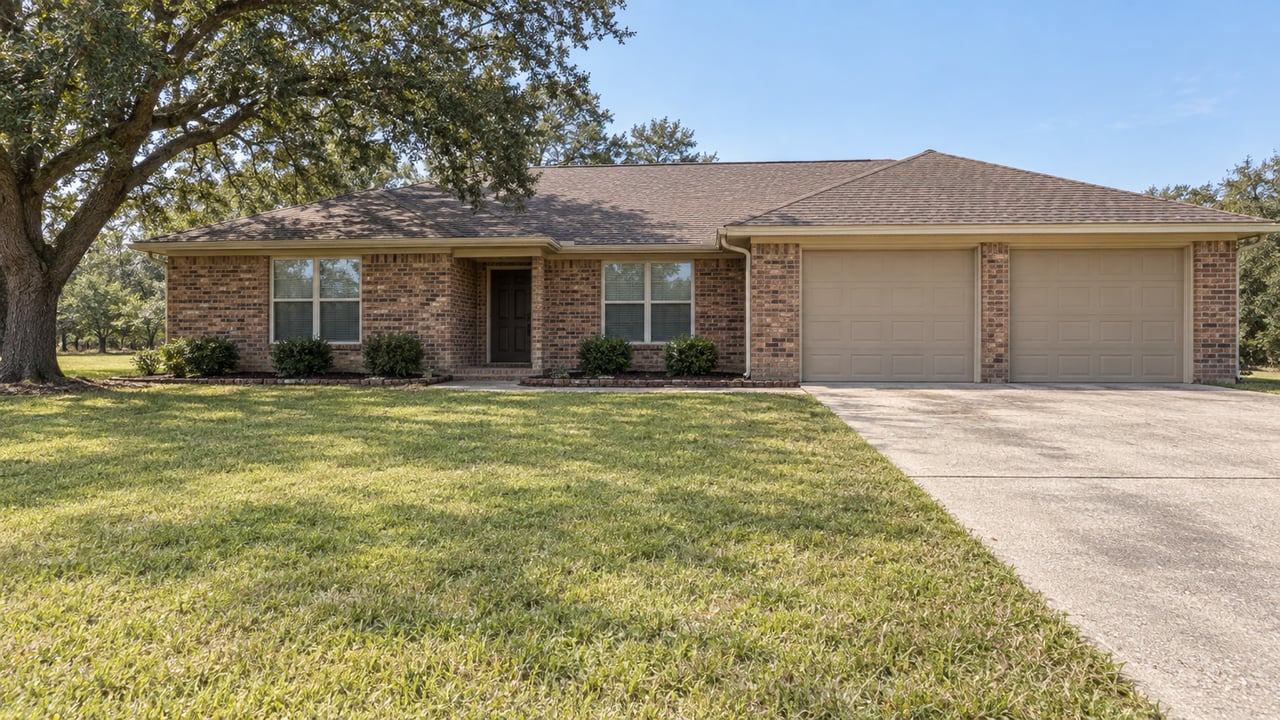 One-story russet brick home behind a broad lawn, with a mature live oak and a concrete driveway leading to two tan garage doors.
