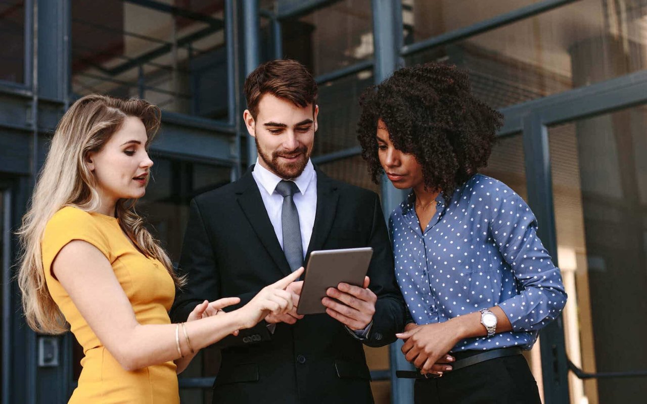 Three professionals review a tablet outside a modern glass-fronted building.
