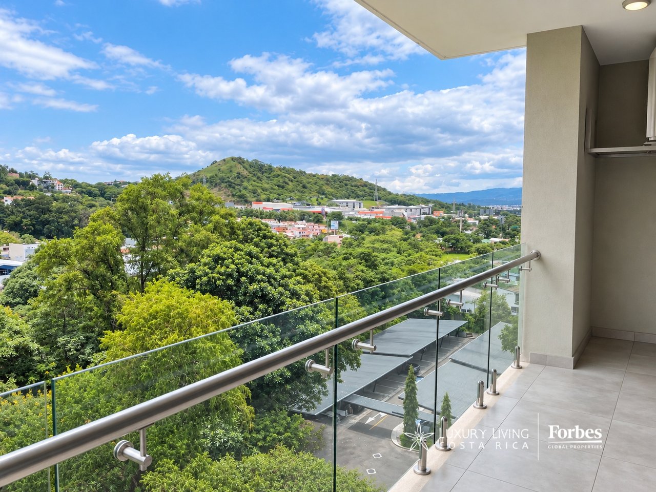 Apartment balcony overlooks leafy hills and a neighborhood, with a glass railing in the foreground.