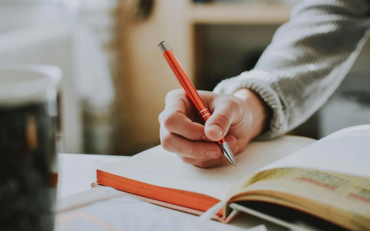 A person writes in a gridded notebook with a red pen beside an open book.