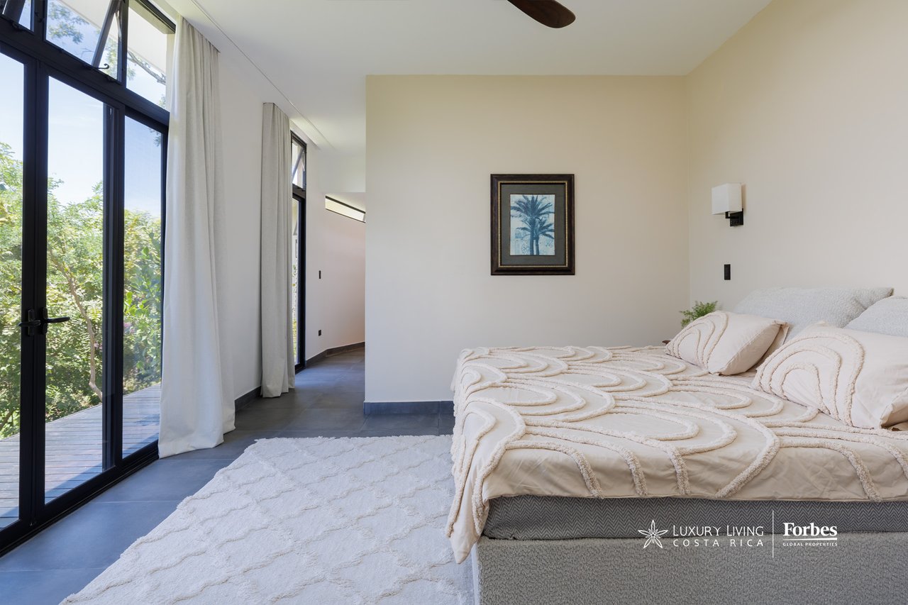 Modern bedroom with textured cream bedding and tall glass doors opening toward leafy greenery.