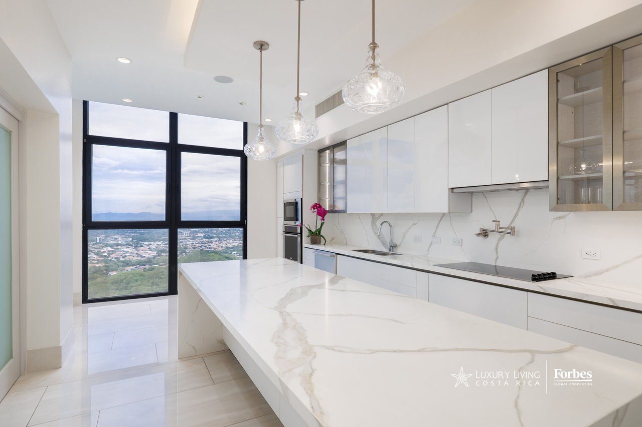 Modern white kitchen with a large marble-look island and floor-to-ceiling city-view windows.