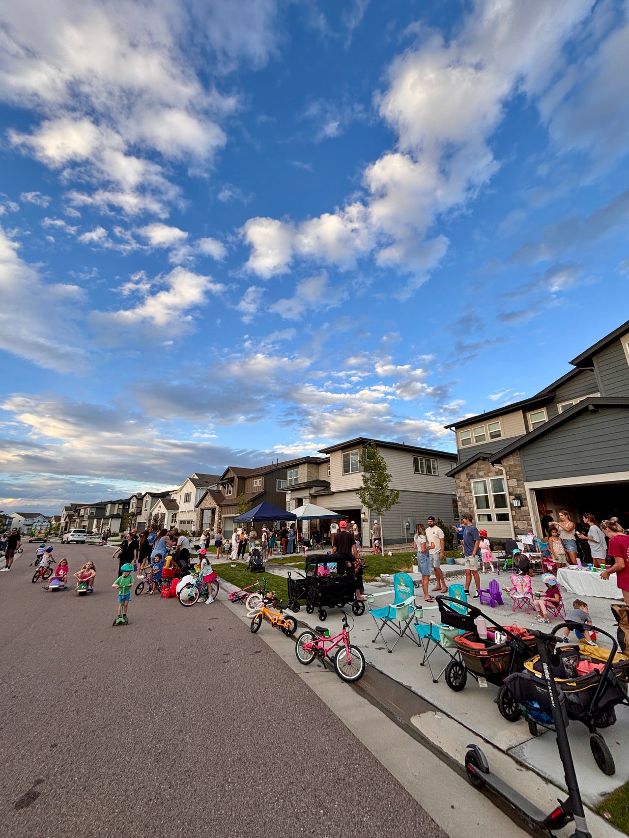 Neighbors gather on a residential street as children ride bikes and scooters beside suburban homes.