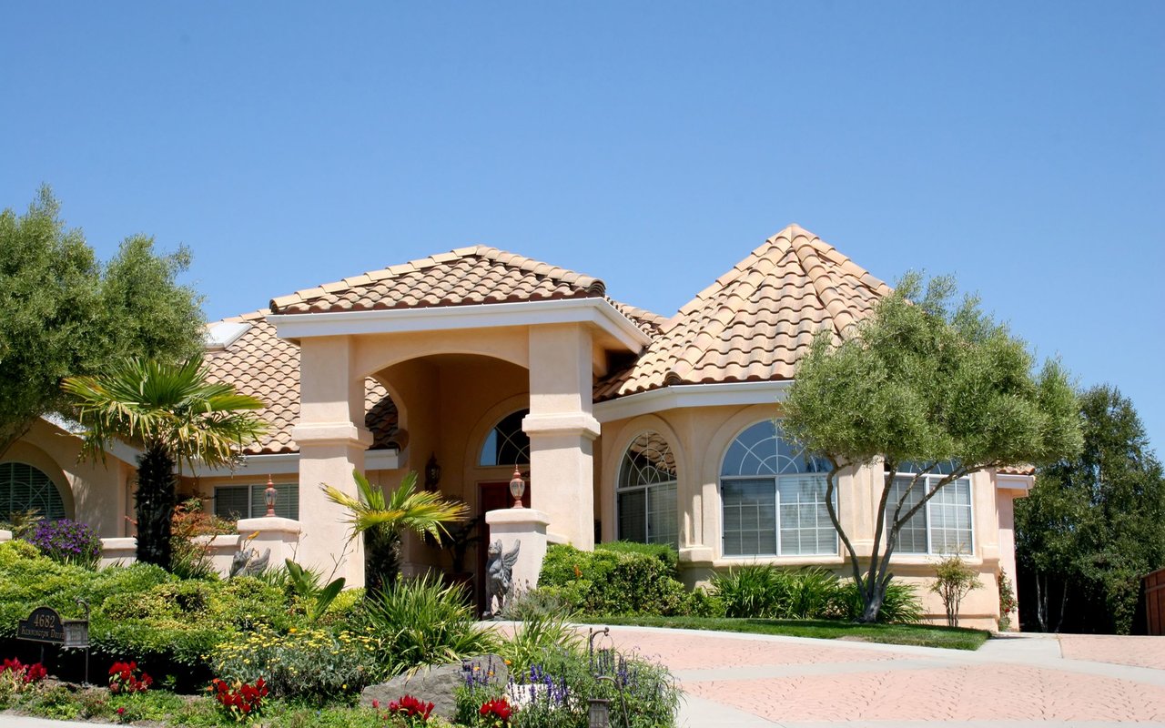 Single-story stucco house with terracotta roof tiles, arched entry, and a landscaped driveway.
