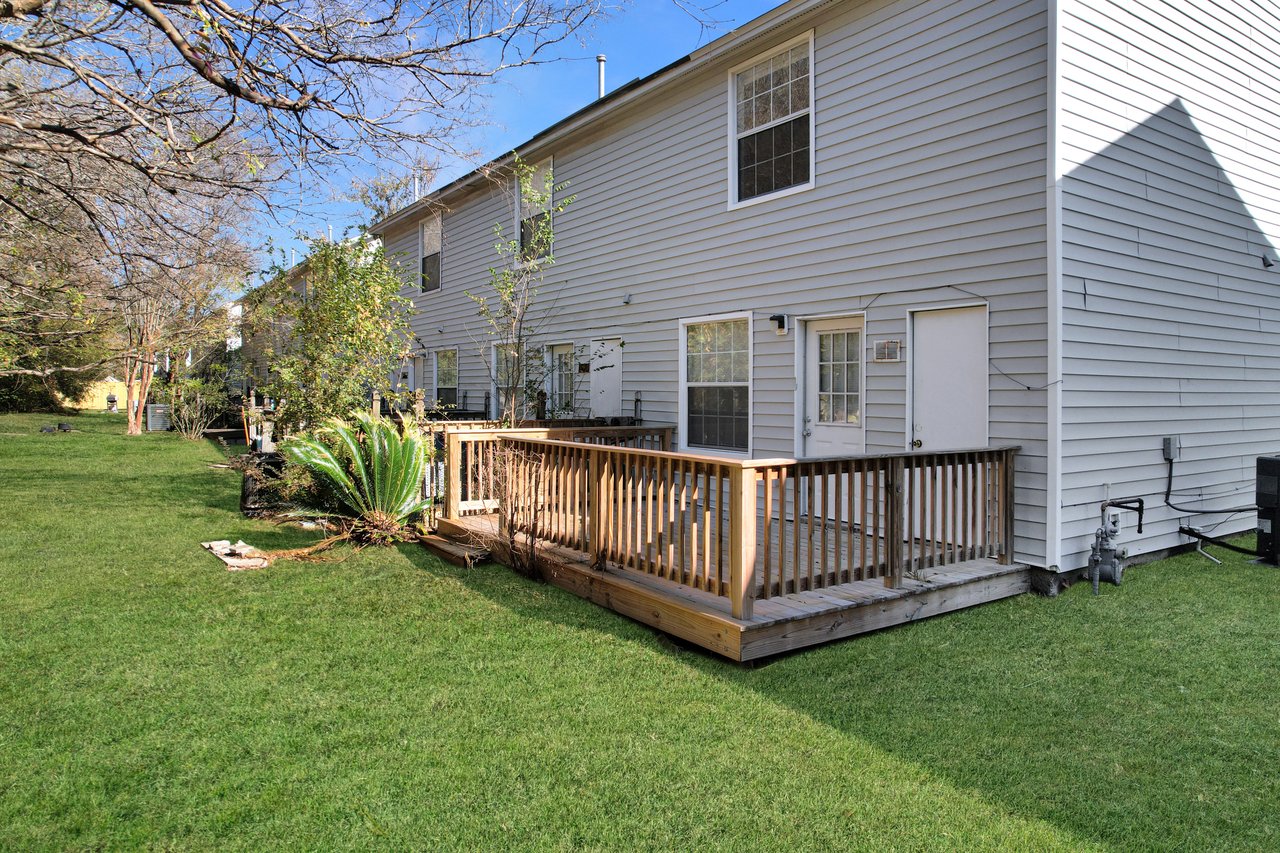 Townhouse exterior with white siding, a small wooden deck, and grassy yard. A few trees and shrubs line the area, evoking a peaceful suburban setting.