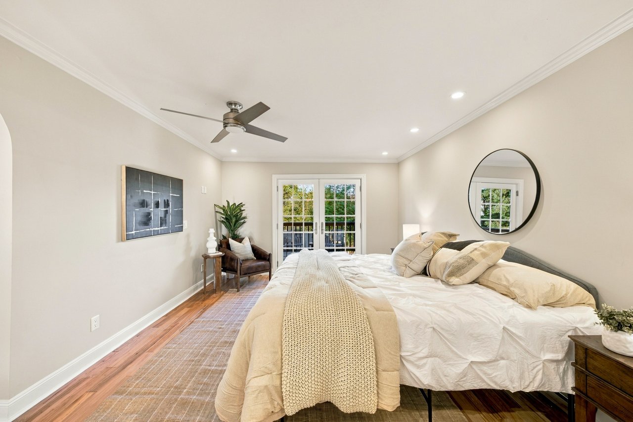 Staged bedroom with a large white bed, round mirror, ceiling fan, and French doors to a balcony.