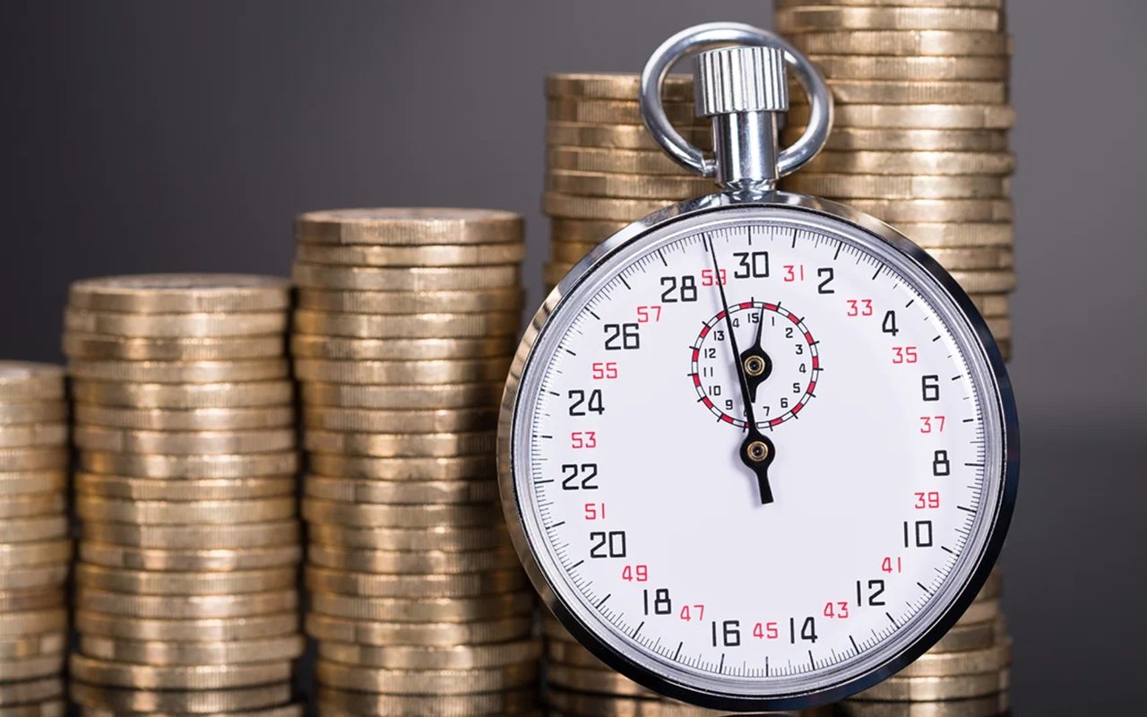 Silver stopwatch in front of rising stacks of gold-colored coins.