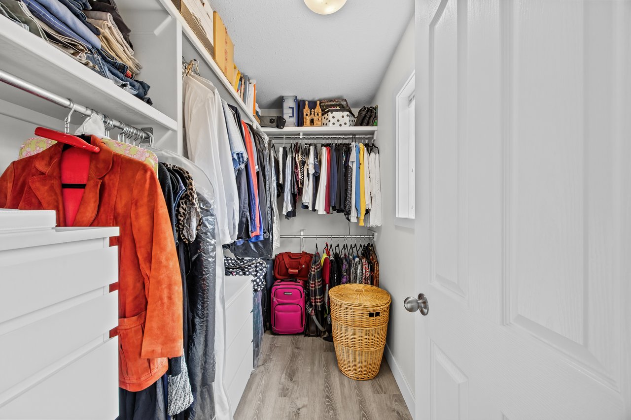 Walk-in closet with built-in white shelving, hanging clothes, pink suitcases, and a wicker hamper.