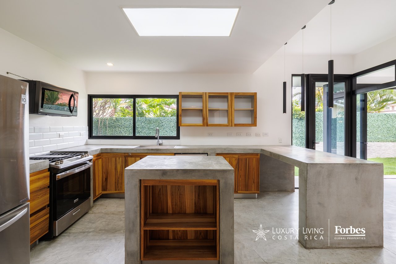 Modern kitchen with a concrete island, wood cabinetry, and windows overlooking a garden.