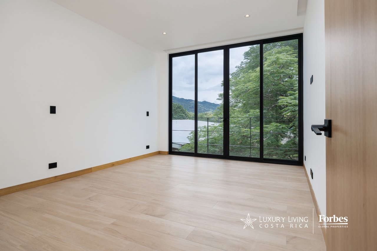 Vacant modern room with tall glass doors, leafy trees, and mountains beyond.