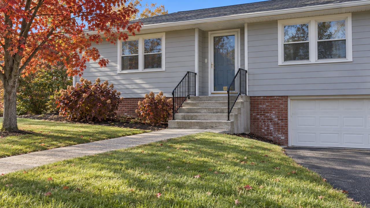 Gray-sided raised-ranch home with a blue entry door, concrete steps, a broad lawn, and red-orange autumn leaves.