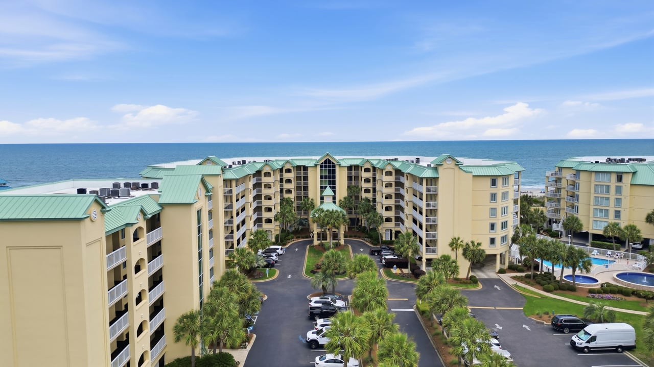 Beachfront condominium buildings with green roofs, palm trees, and a parking courtyard beside the ocean.