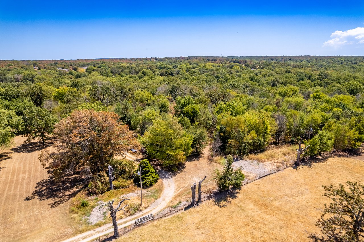 Aerial view of wooded land bordered by dry fields, with a dirt road and fencing.