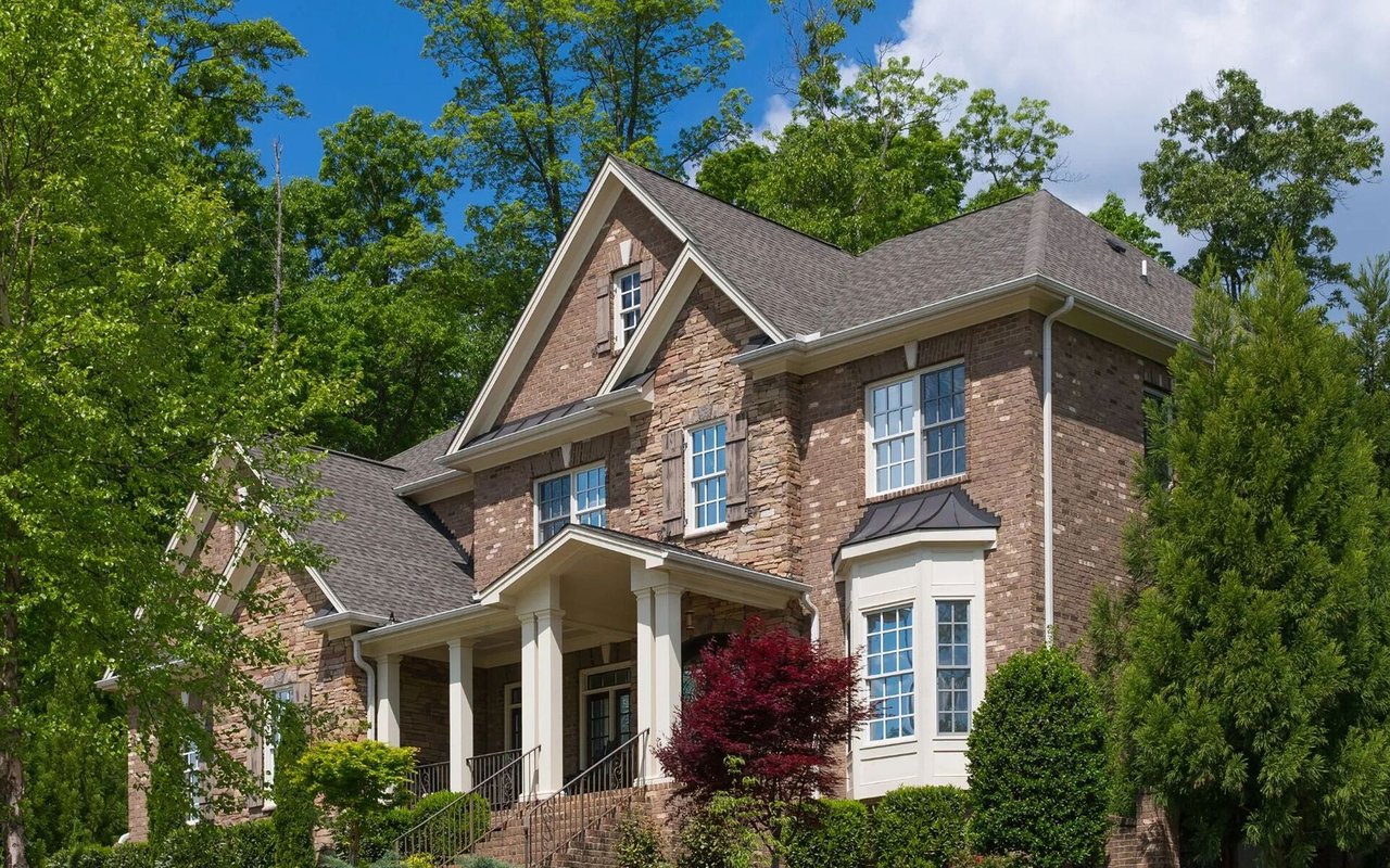 Two-story brick and stone house with a columned porch among mature trees.