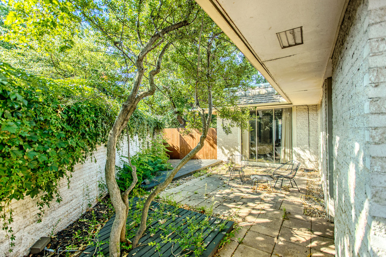 Shaded paved patio with outdoor chairs, leafy trees, and a sliding glass door.