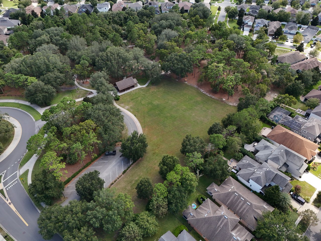 Aerial view of a tree-lined neighborhood park with walking paths, a covered pavilion, and surrounding homes.