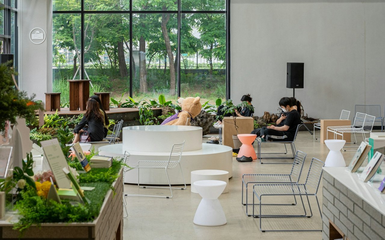 Commercial lounge with modern seating, indoor plants, large windows, and people relaxing.