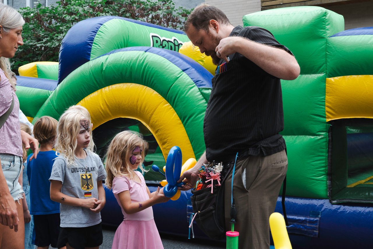 Balloon artist gives children a blue balloon sculpture beside a colorful inflatable bounce house.