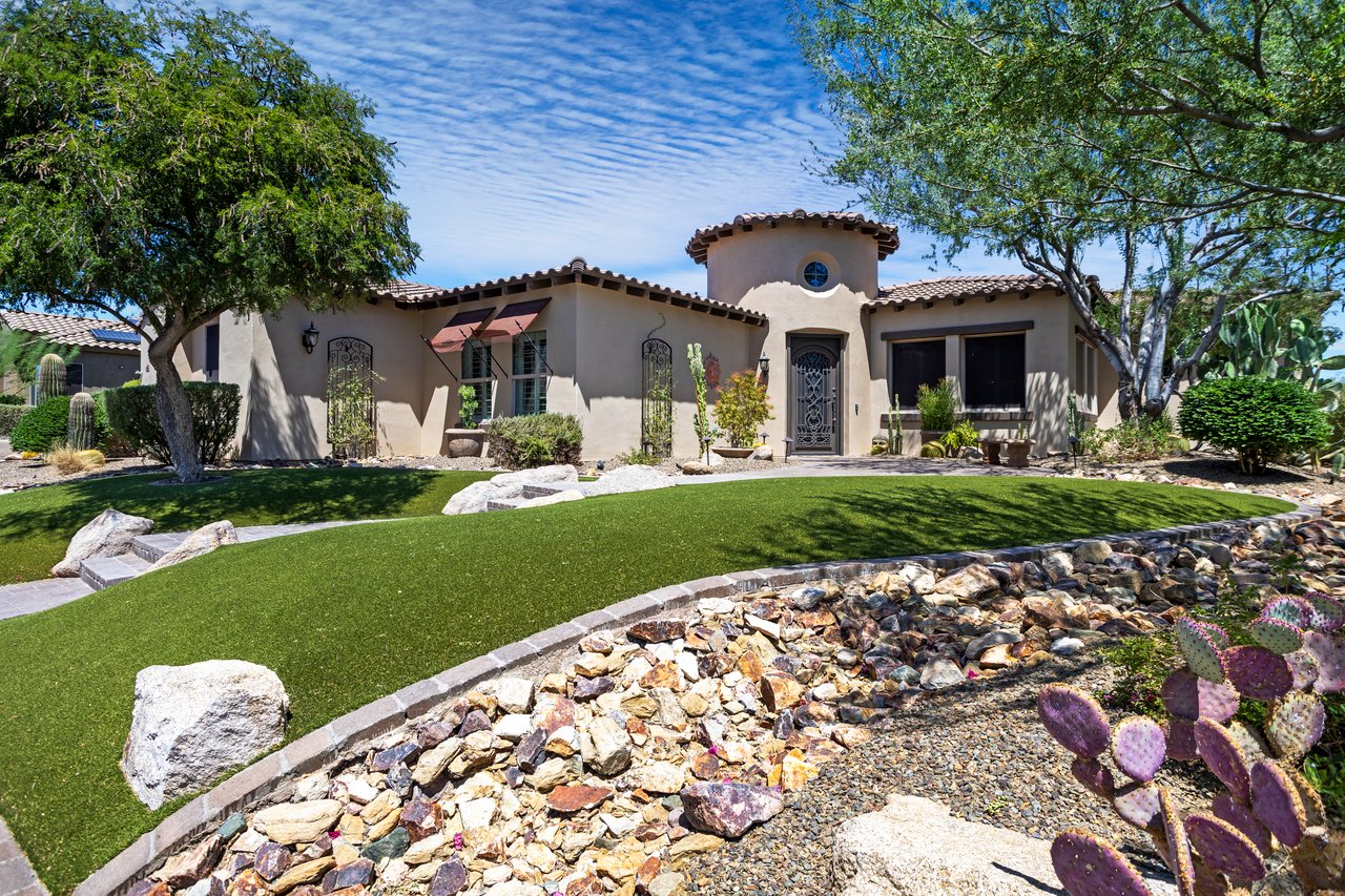 Front exterior of a single-story stucco house with a landscaped yard, desert plants, and a tiled roof under a blue sky.