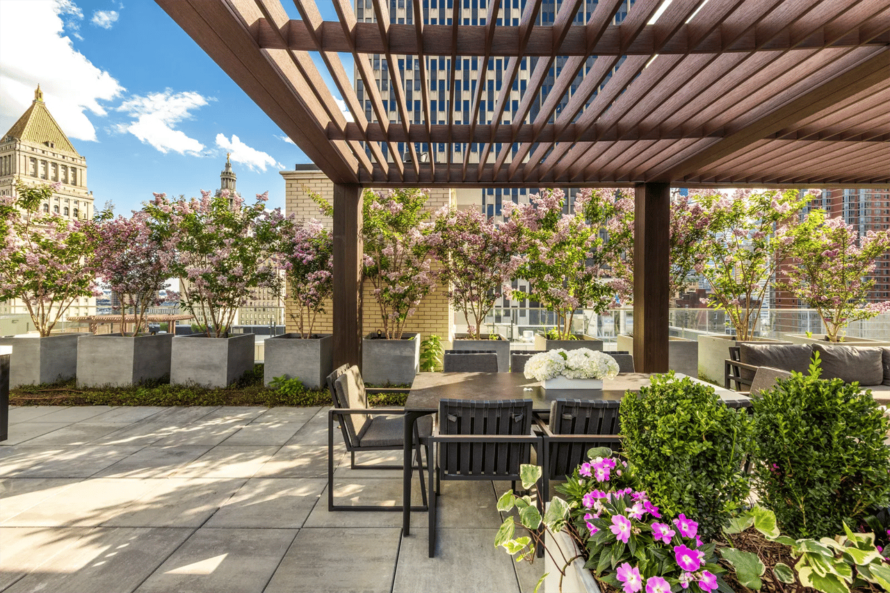 Rooftop terrace with a wood pergola, outdoor seating, and flowering planters overlooking city buildings.