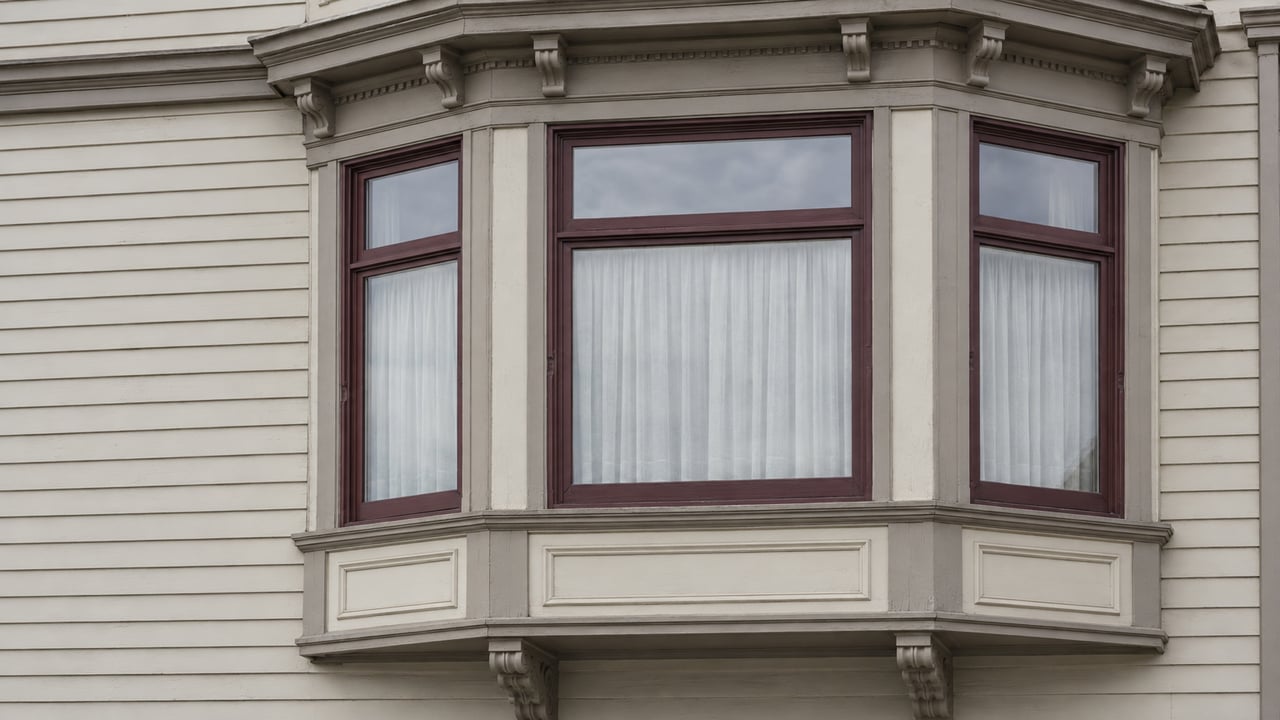 Close upward view of a cream-painted timber bay with burgundy sash windows, carved brackets and softly weathered molding.