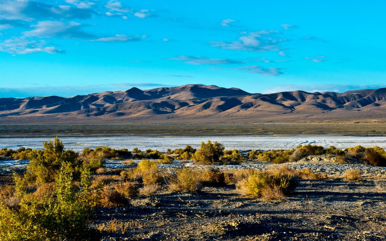 Rocky valley with golden shrubs, a riverbed, and mountain ridges beneath a blue sky.