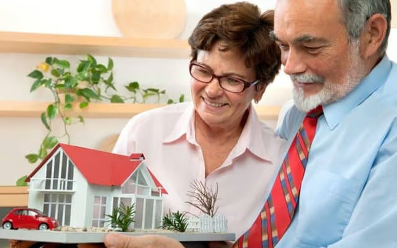 Older couple examining a model house with a red roof and a small red car.