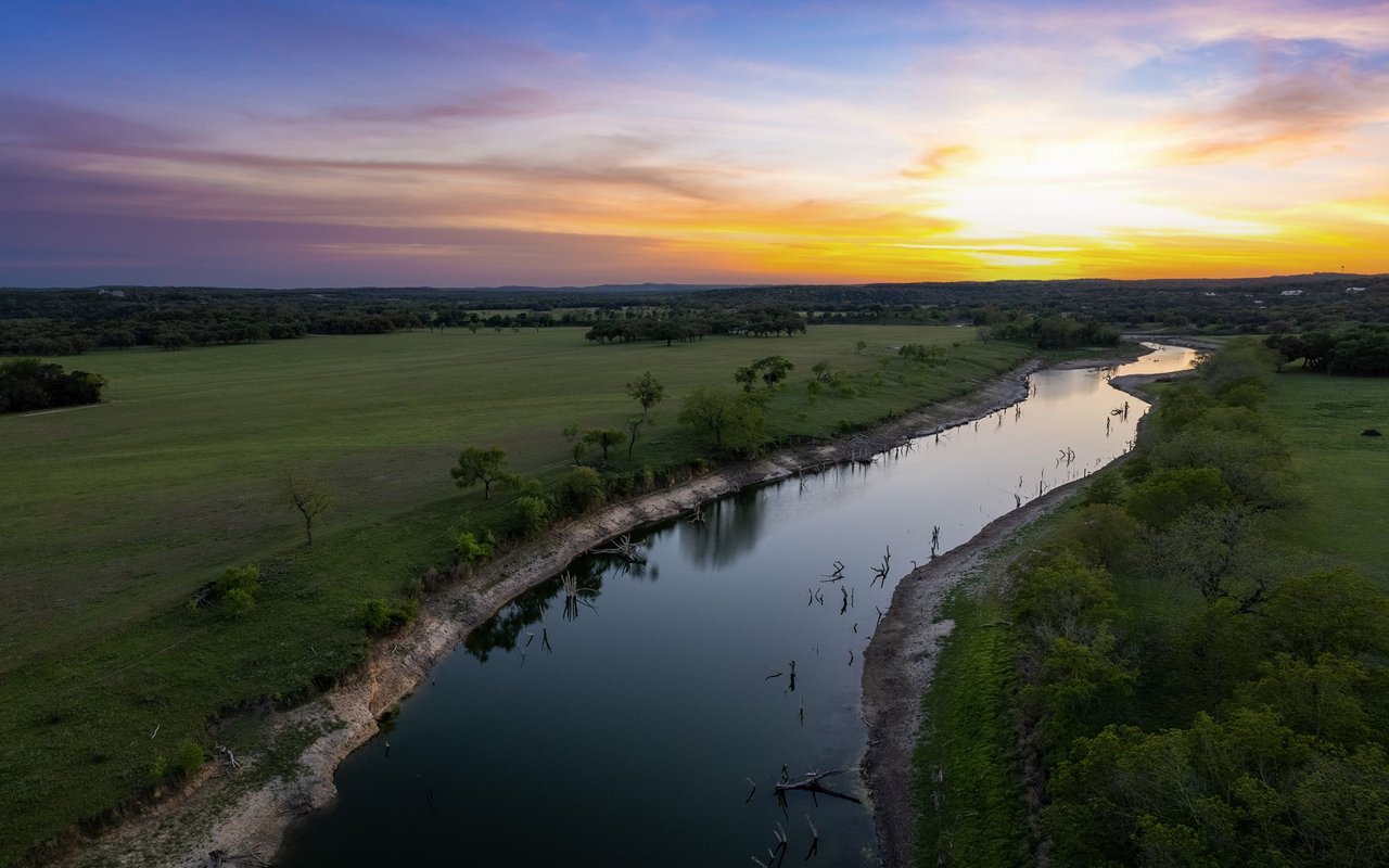 Winding creek through open pasture and wooded land beneath a colorful sunset sky.
