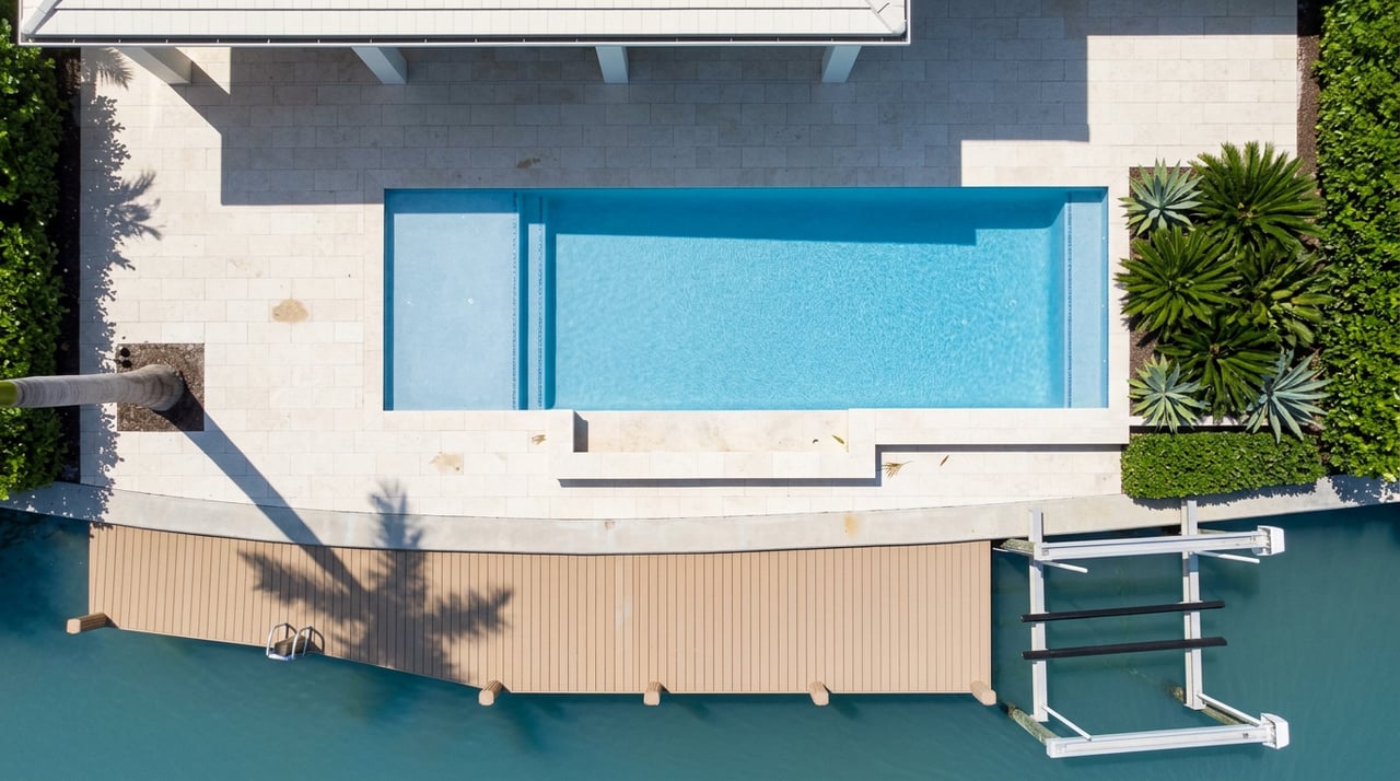 Overhead view of a tiled pool, pale stone terrace, tan dock, and boat lift beside blue-green canal water.